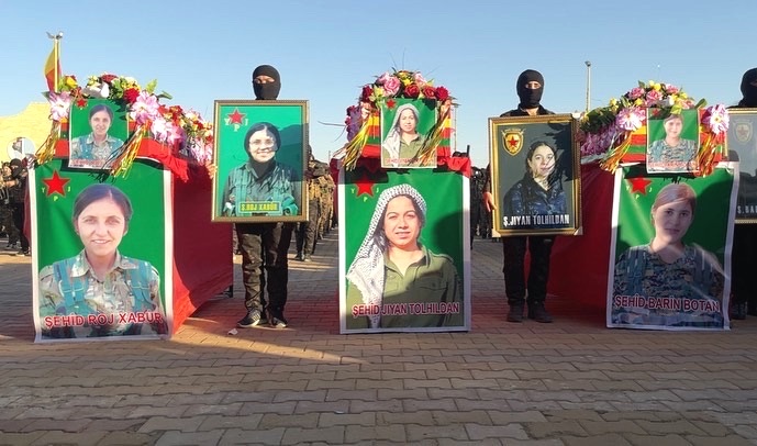 Große Bilder der drei getöteten Frauen werden auf der Straße gezeigt. Im Hintergrund stehen Kämpferinnen und Kämpfer in Militärkleidung. Große Bilder der drei getöteten Frauen werden auf der Straße gezeigt. Im Hintergrund stehen Kämpferinnen und Kämpfer in Militärkleidung.