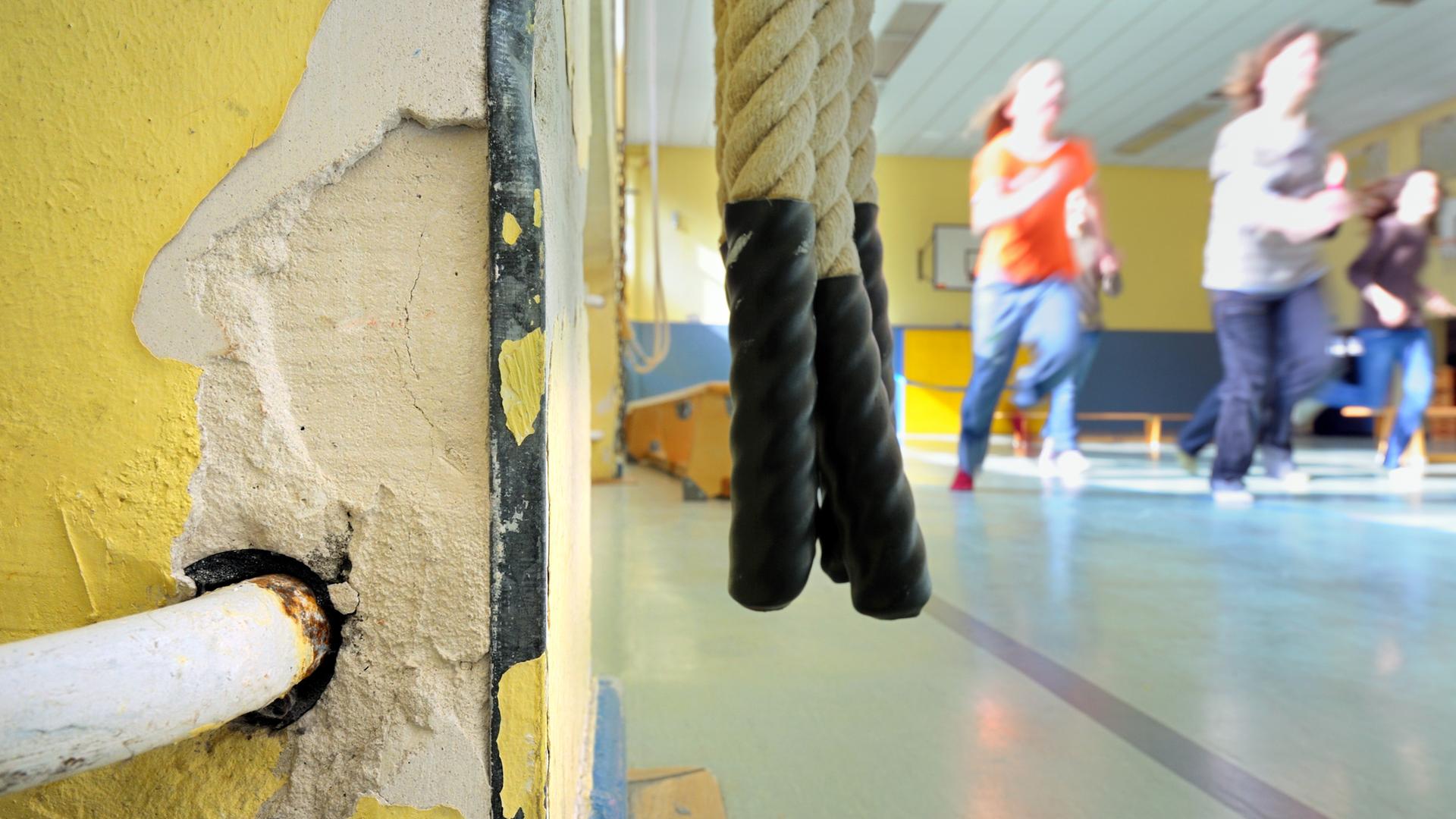 Kinder laufen in einer Turnhalle an der Zuleitung eines Heizkörpers vorbei, in dessen Umgebung bereits der Putz abgebröckelt ist. Kinder laufen in einer Turnhalle an der Zuleitung eines Heizkörpers vorbei, in dessen Umgebung bereits der Putz abgebröckelt ist.