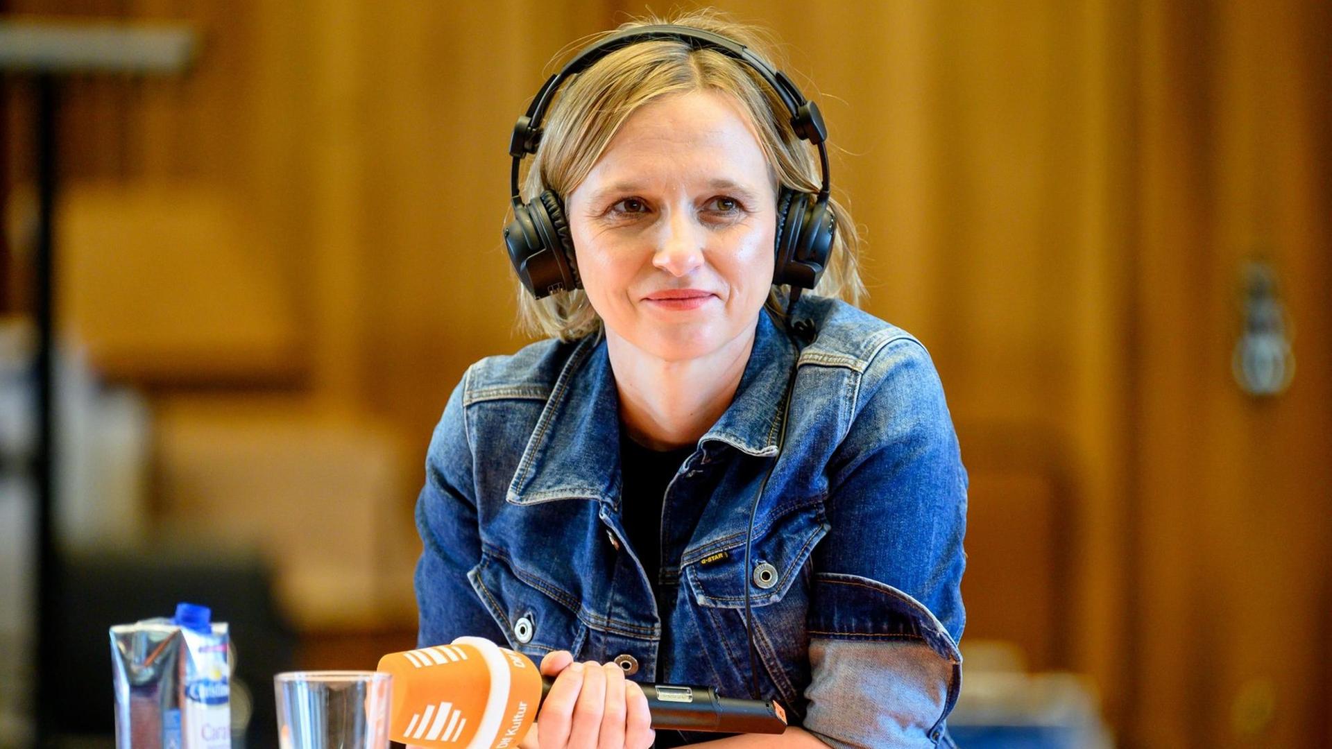 Das Bild zeigt eine junge Frau mit Mikrofon und Kopfhörern, die an einem Tisch sitzt und lächelt.
