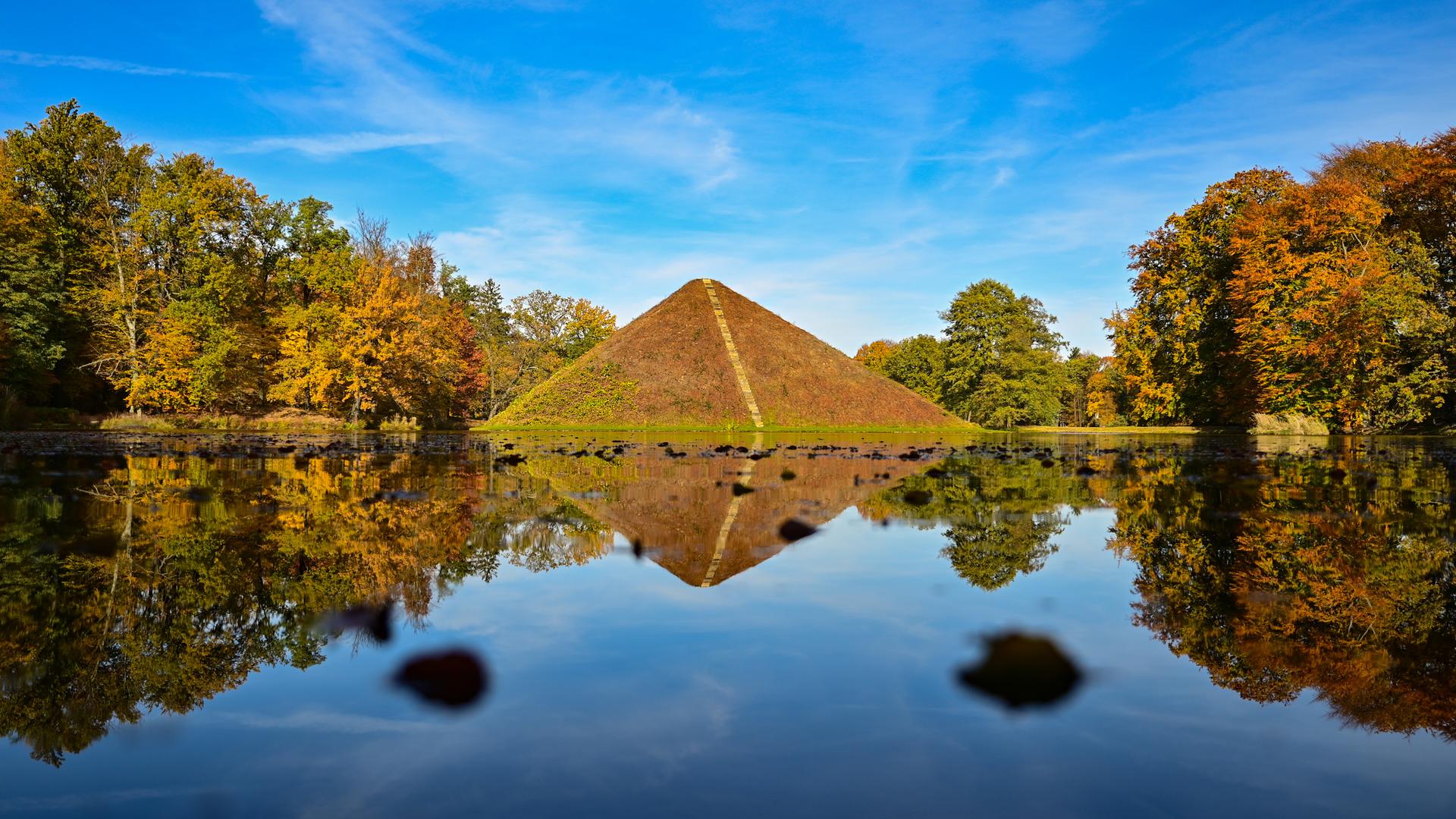 Eine bepflanzte Steinpyramide in einem See, der von herbstlich gefärbten Bäumen umstanden ist Eine bepflanzte Steinpyramide in einem See, der von herbstlich gefärbten Bäumen umstanden ist