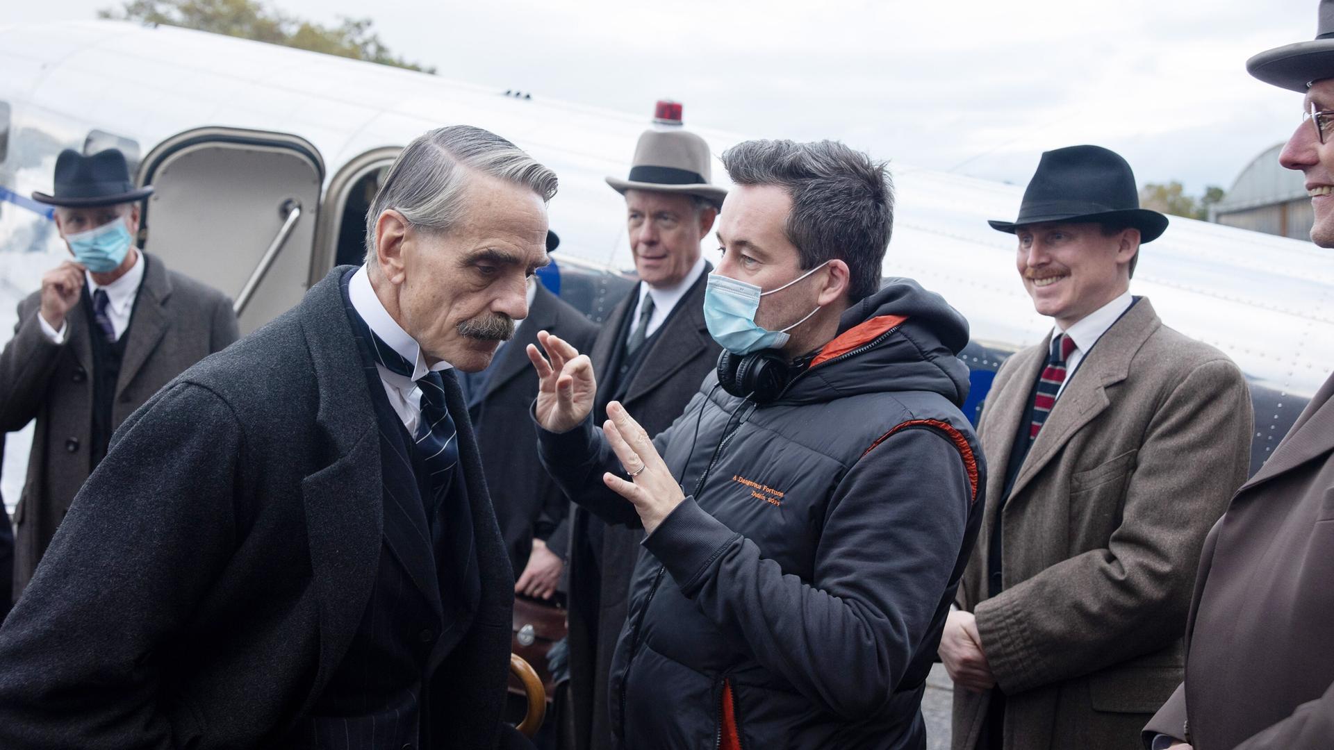 Am Set von "MUNICH: THE EDGE OF WAR" gibt Regisseur Christian Schwochow Hauptdarsteller Jeremy Irons Anweisungen. Schwochow trägt eine Schutzmaske, die anderen im Bild tragen Anzüge und Hüte im Stil der 1930er-Jahre. Am Set von "MUNICH: THE EDGE OF WAR" gibt Regisseur Christian Schwochow Hauptdarsteller Jeremy Irons Anweisungen. Schwochow trägt eine Schutzmaske, die anderen im Bild tragen Anzüge und Hüte im Stil der 1930er-Jahre.