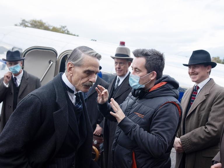 Am Set von "MUNICH: THE EDGE OF WAR" gibt Regisseur Christian Schwochow Hauptdarsteller Jeremy Irons Anweisungen. Schwochow trägt eine Schutzmaske, die anderen im Bild tragen Anzüge und Hüte im Stil der 1930er-Jahre.