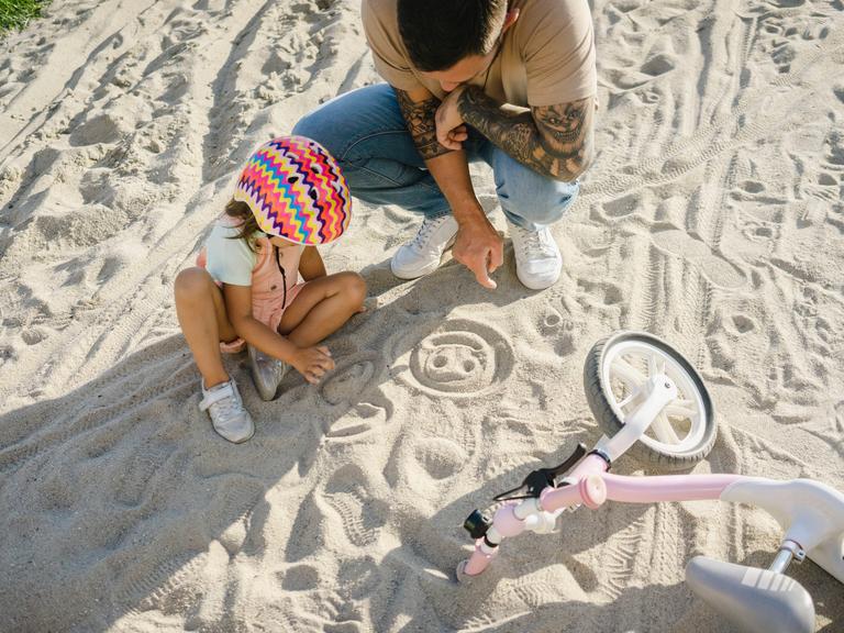 Vater und Tochter sitzen am Strand und malen lachende Gesichter in den Sand.