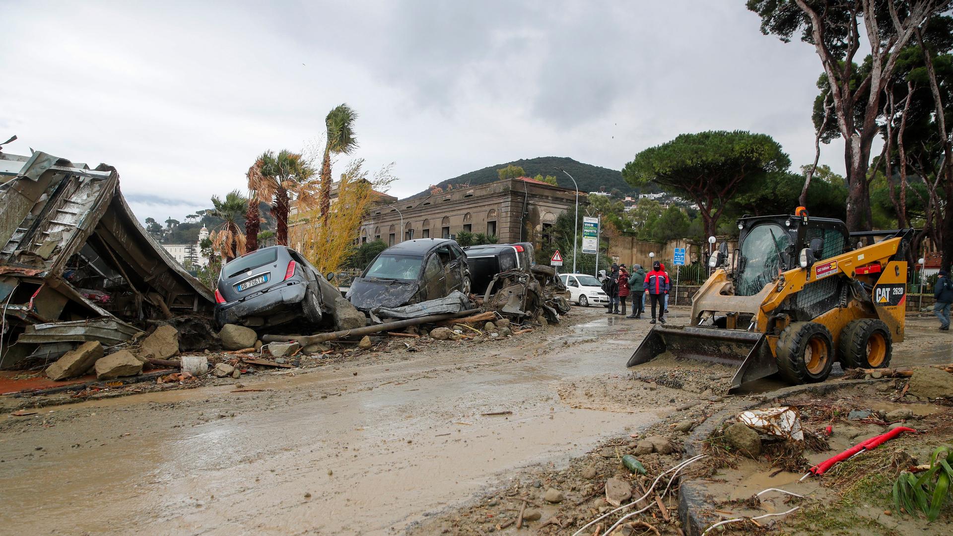 Eine Straße im Schlamm, an deren Rändern Räumfahrzeuge, kaputte Autos und ein eingestürztes Gebäude. Eine Straße im Schlamm, an deren Rändern Räumfahrzeuge, kaputte Autos und ein eingestürztes Gebäude.