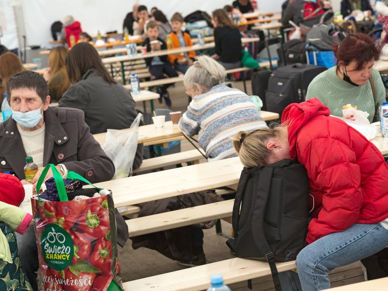 Erschöpfte ukrainische Flüchtlinge sitzen im Willkommenszelt am Berliner Hauptbahnhof auf Holzbänken. 