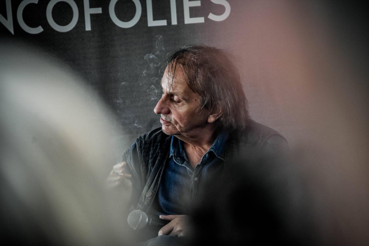 Michel Houellebecq sitzt mit genervtem Gesichtsausdruck auf einem Podium Michel Houellebecq sitzt mit genervtem Gesichtsausdruck auf einem Podium