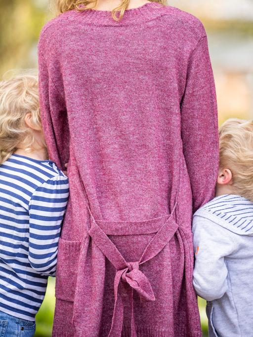 Zu sehen ist eine Frau von hinten, an die sich jeweils ein Junge und ein Mädchen anschmiegen. Vermutlich Sohn und Tochter. 