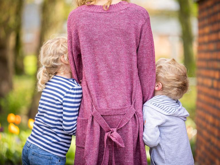 Zu sehen ist eine Frau von hinten, an die sich jeweils ein Junge und ein Mädchen anschmiegen. Vermutlich Sohn und Tochter. 