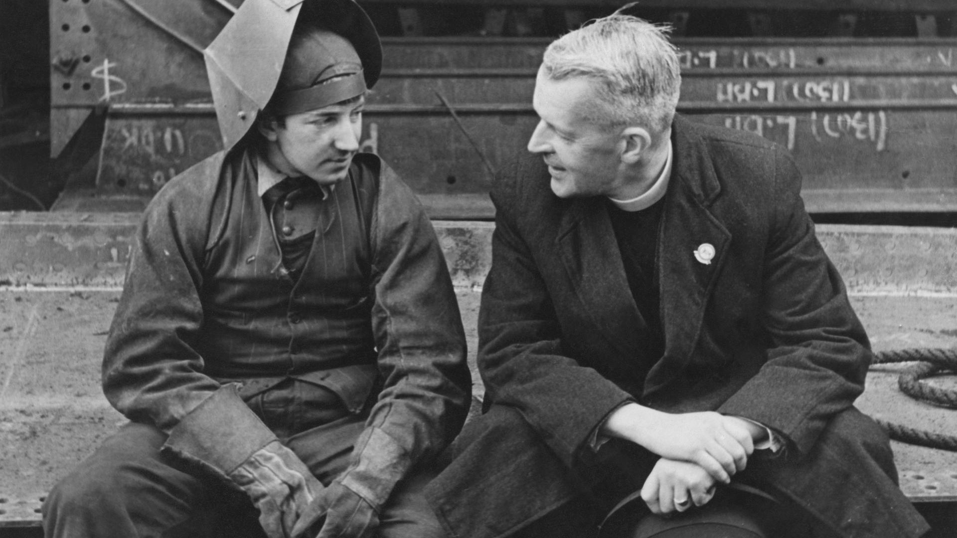 Historische Schwarzweißaufnahme eines Schweißers und eines Priesters, die sich sitzend vor einer Industrieszenerie, unterhalten.