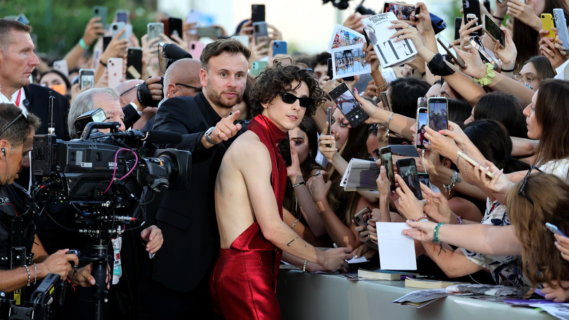 Timothée Chalamet steht vor einer Absperrung und signiert Autogramme, während auf der anderen Seite des Zauns dutzende Fans begeistert ihre Smartphones auf ihn richten. Timothée Chalamet steht vor einer Absperrung und signiert Autogramme, während auf der anderen Seite des Zauns dutzende Fans begeistert ihre Smartphones auf ihn richten.