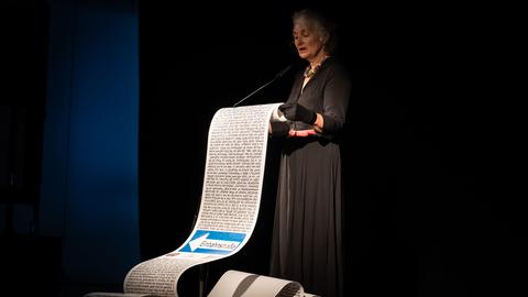 Eine Frau im schwarzen Kleid und mit grauen Haaren steht auf einer Bühne und liest aus einer überdimensionalen Schriftrolle vor, die sie in ihren Händen hält.