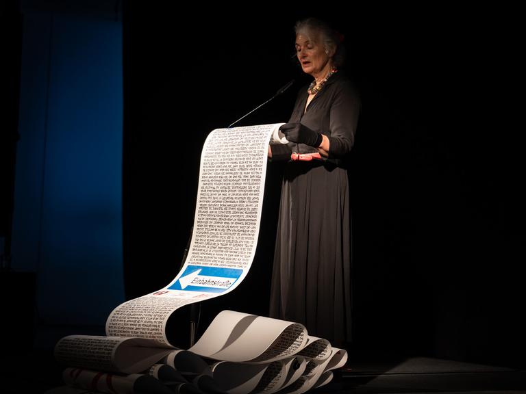 Eine Frau im schwarzen Kleid und mit grauen Haaren steht auf einer Bühne und liest aus einer überdimensionalen Schriftrolle vor, die sie in ihren Händen hält.