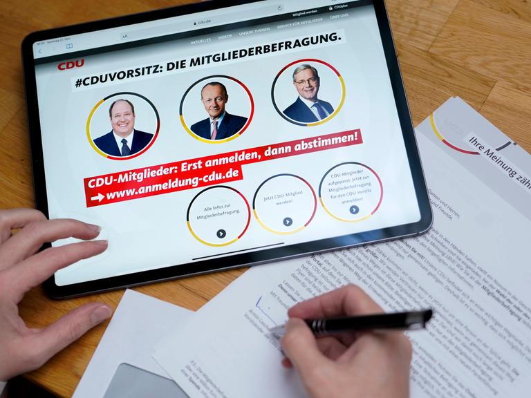 Auf einem Tablet sind in runde Bilder von drei Männern zu sehen: Helge Braun, Friedrich Merz und Norbert Röttgen. Darüber steht "CDU Vorsitz Mitgliederbefragung". Eine Hand hält einen Stift über einen Zettel. 