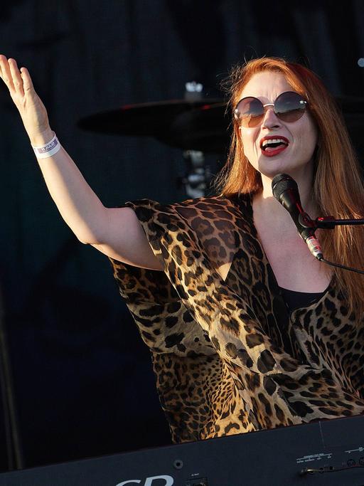 Die walisische Sängerin Judith Owen ist bei einem Konzert mit runder Sonnenbrille auf der Bühne hinter einem Mikrofon zu sehen und hebt die Arme in die Höhe. 