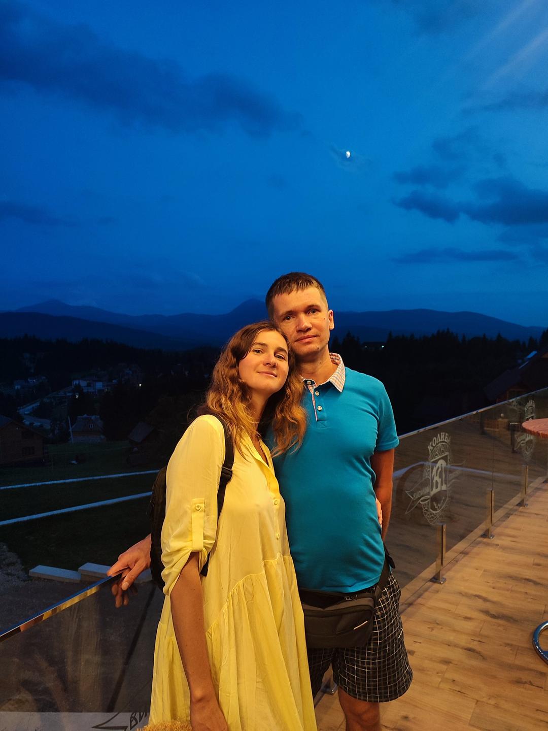 Ein junges Paar steht Arm in Arm bei Abendlicht auf einer Terrasse. Im Hintergrund sind die Berge zu sehen.