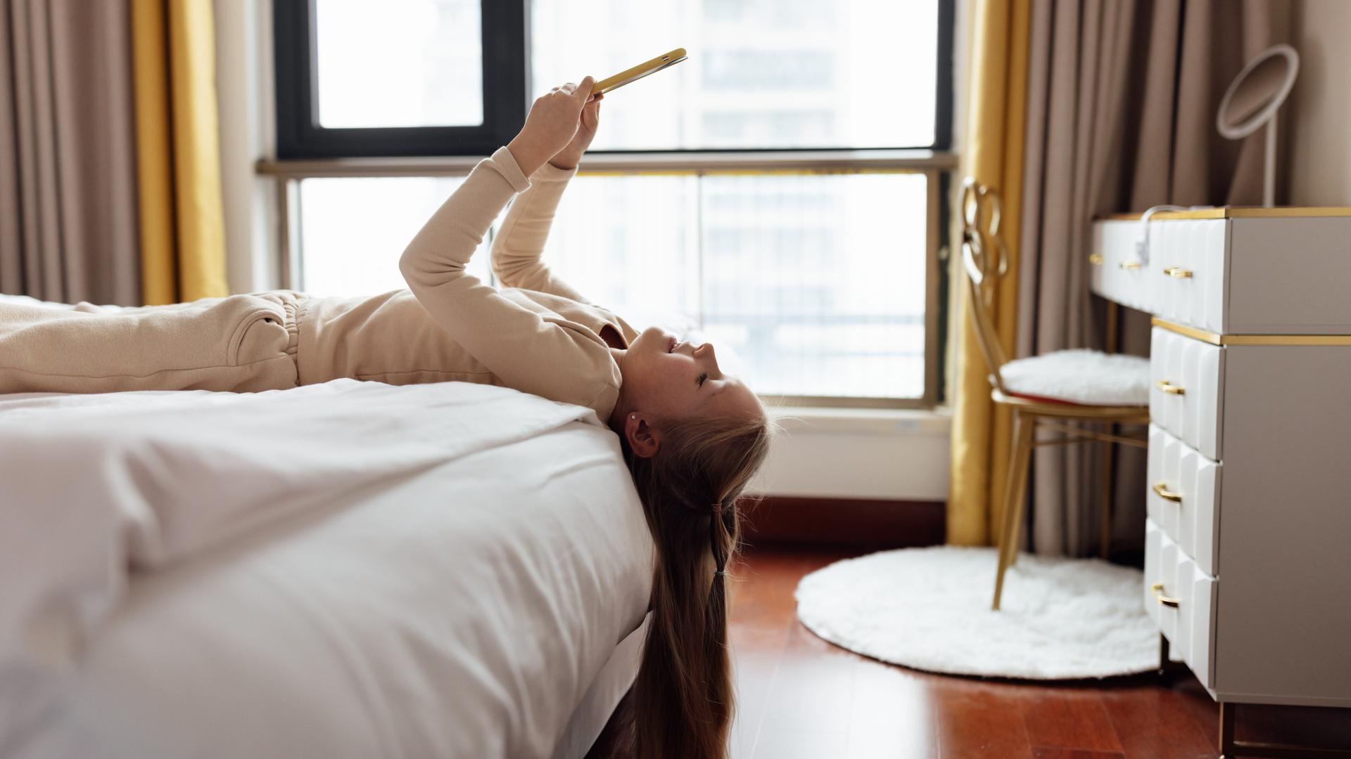 Ein Mädchen liegt auf dem Rücken auf einem Hotelbett, das Smartphone über das Gesicht gehoben. Die Haare hängen vorn am Bett herunter. Ein Mädchen liegt auf dem Rücken auf einem Hotelbett, das Smartphone über das Gesicht gehoben. Die Haare hängen vorn am Bett herunter.