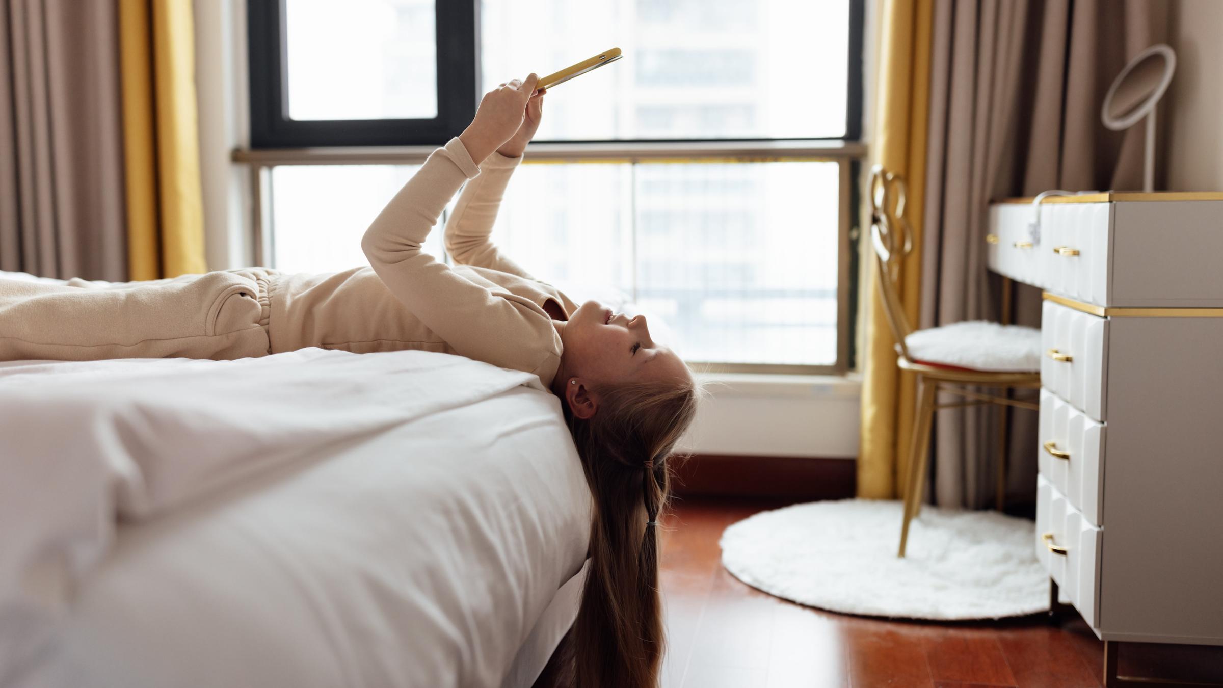 Ein Mädchen liegt auf dem Rücken auf einem Hotelbett, das Smartphone über das Gesicht gehoben. Die Haare hängen vorn am Bett herunter.