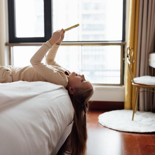 Ein Mädchen liegt auf dem Rücken auf einem Hotelbett, das Smartphone über das Gesicht gehoben. Die Haare hängen vorn am Bett herunter.
