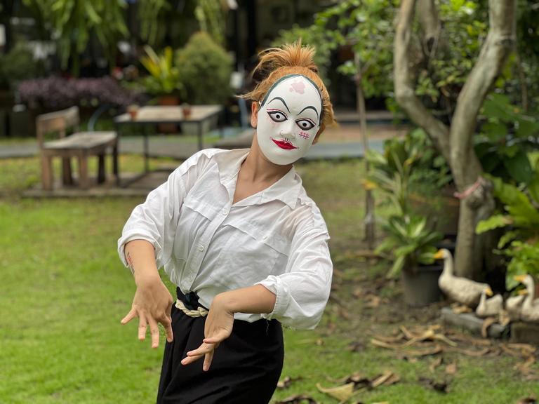 Eine junge Frau bewegt sich - auf dem Gesicht weiße bemalte Papp-Maske - rhythmisch durch einen Garten.