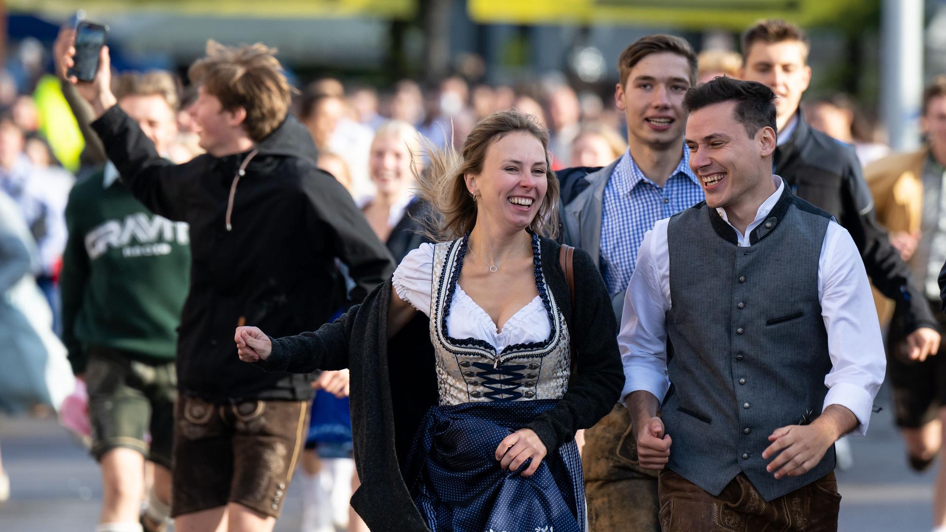 Die ersten Wiesnbesucher rennen nach dem Einlass zu den Festzelten. Im Vordergrund ist eine lachende, junge Frau im Dirndl zu sehen, rechts neben ihr ein junger Mann in Lederhose und Weste über weißem Hemd. Auch die Menschen im Hintergrund strahlen voller Vorfreude auf den Wiesnbesuch. Links im Bild richtet ein junger Mann in Lederhose und schwarzem Hoodie ein Handy auf sich. Die ersten Wiesnbesucher rennen nach dem Einlass zu den Festzelten. Im Vordergrund ist eine lachende, junge Frau im Dirndl zu sehen, rechts neben ihr ein junger Mann in Lederhose und Weste über weißem Hemd. Auch die Menschen im Hintergrund strahlen voller Vorfreude auf den Wiesnbesuch. Links im Bild richtet ein junger Mann in Lederhose und schwarzem Hoodie ein Handy auf sich.