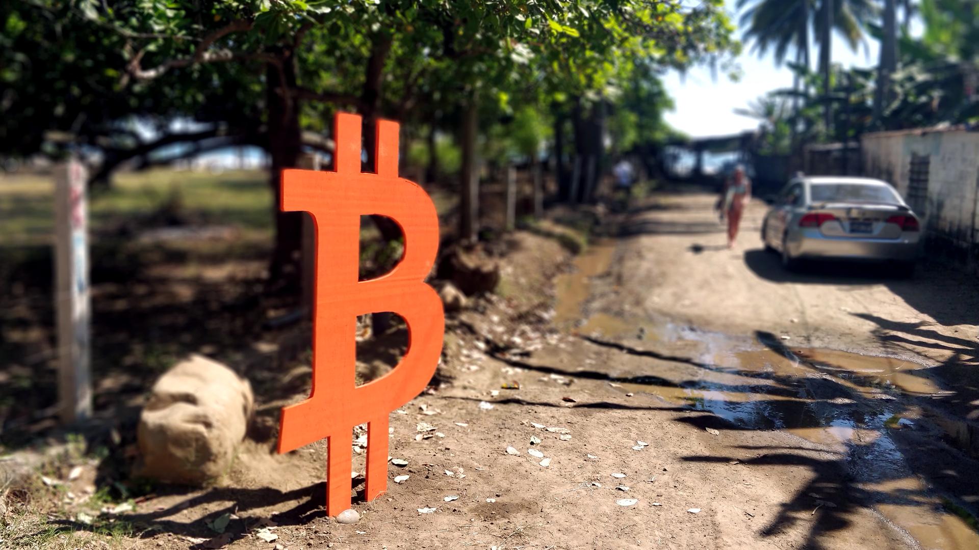 Ein mannshohes Bitcoin-Zeichen steht an einer Straße, die zum Meer führt. Ein mannshohes Bitcoin-Zeichen steht an einer Straße, die zum Meer führt.