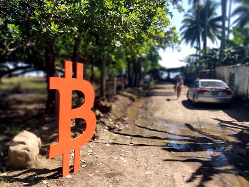 Ein mannshohes Bitcoin-Zeichen steht an einer Straße, die zum Meer führt. 
