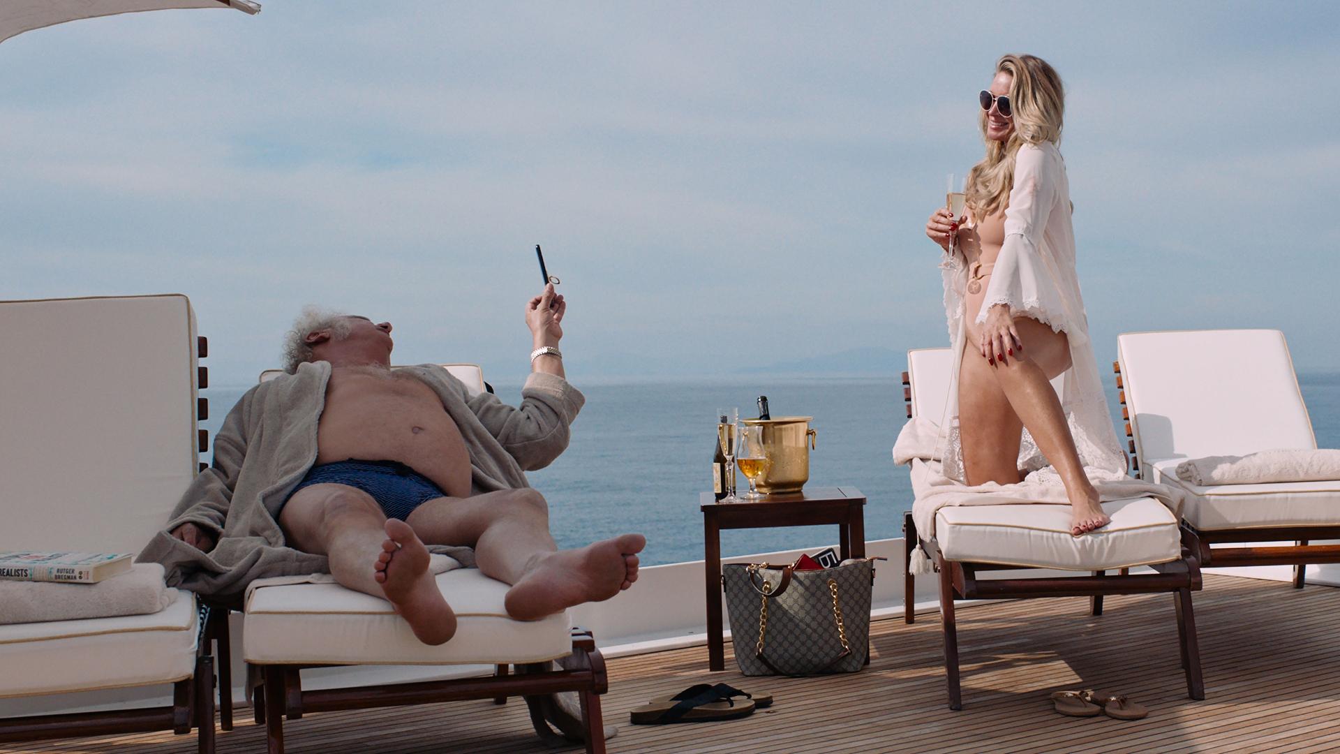 Filmstill aus "Triangle of Sadness": Eine blonde und leicht bekleidete Frau schäkert an Bord einer Luxusjacht mit einem älteren Mann.