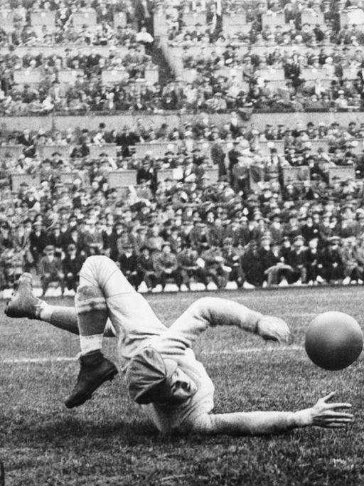 Schwarzweißfoto von einem historischen Fußballspiel. Ein Torhüter liegt auf dem Fußballfeld und fängt einen Fußball. 