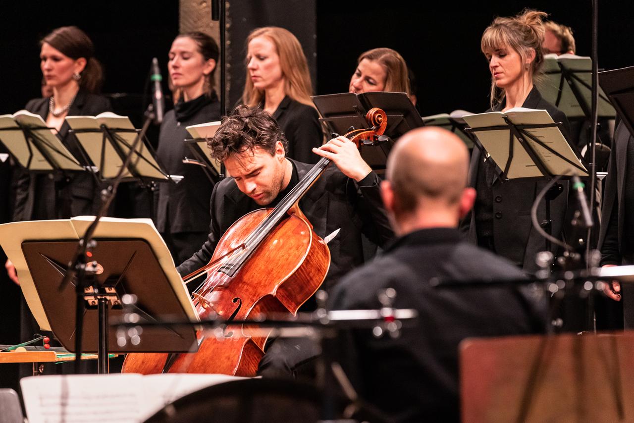 Der Cellist Leonard Elschenbroich spielt engagiert sein Instrument, während er von Instrumenten und Sängern umringt ist. Der Cellist Leonard Elschenbroich spielt engagiert sein Instrument, während er von Instrumenten und Sängern umringt ist.