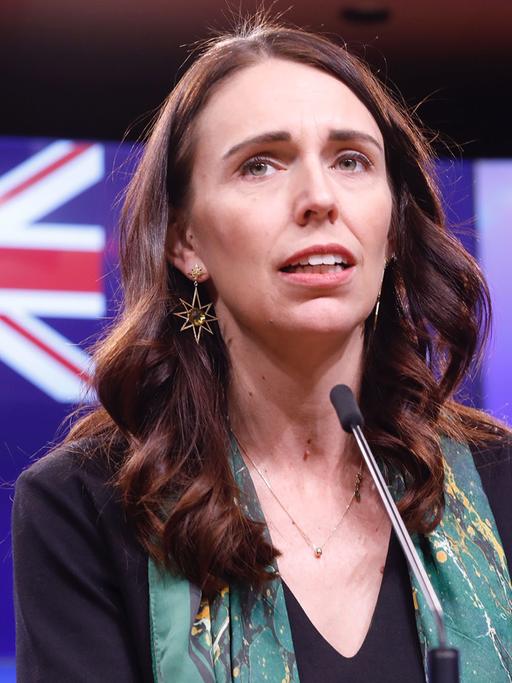 Das Bild zeigt eine schlanke, dunkelhaarige Frau mit langen Haaren. Sie spricht in ein Mikrofon, im Hintergrund ist die neuseeländische Fahne zu sehen. Die Frau ist Jacinda Ardern, von Oktober 2017 bis Ende Januar 2023 Ministerpräsidentin von Neuseeland.