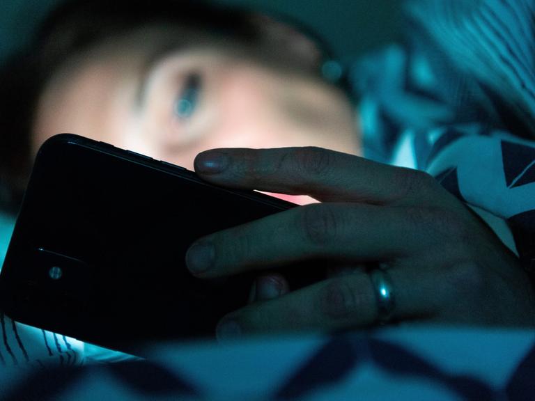 Eine Person liegt im Bett und schaut auf ein Smartphone. 