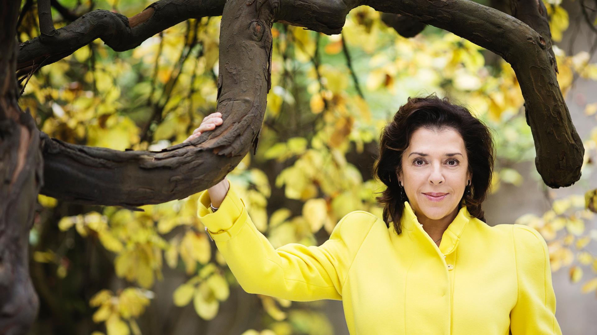 Elena Bashkirova steht in einem gelben Kostüm unter einem gelb belaubten Baum mit markanten, dunklen Ästen. Elena Bashkirova steht in einem gelben Kostüm unter einem gelb belaubten Baum mit markanten, dunklen Ästen.