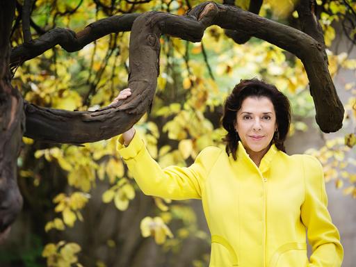 Elena Bashkirova steht in einem gelben Kostüm unter einem gelb belaubten Baum mit markanten, dunklen Ästen.