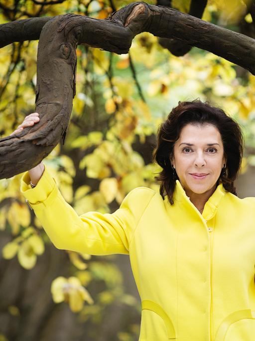 Elena Bashkirova steht in einem gelben Kostüm unter einem gelb belaubten Baum mit markanten, dunklen Ästen.