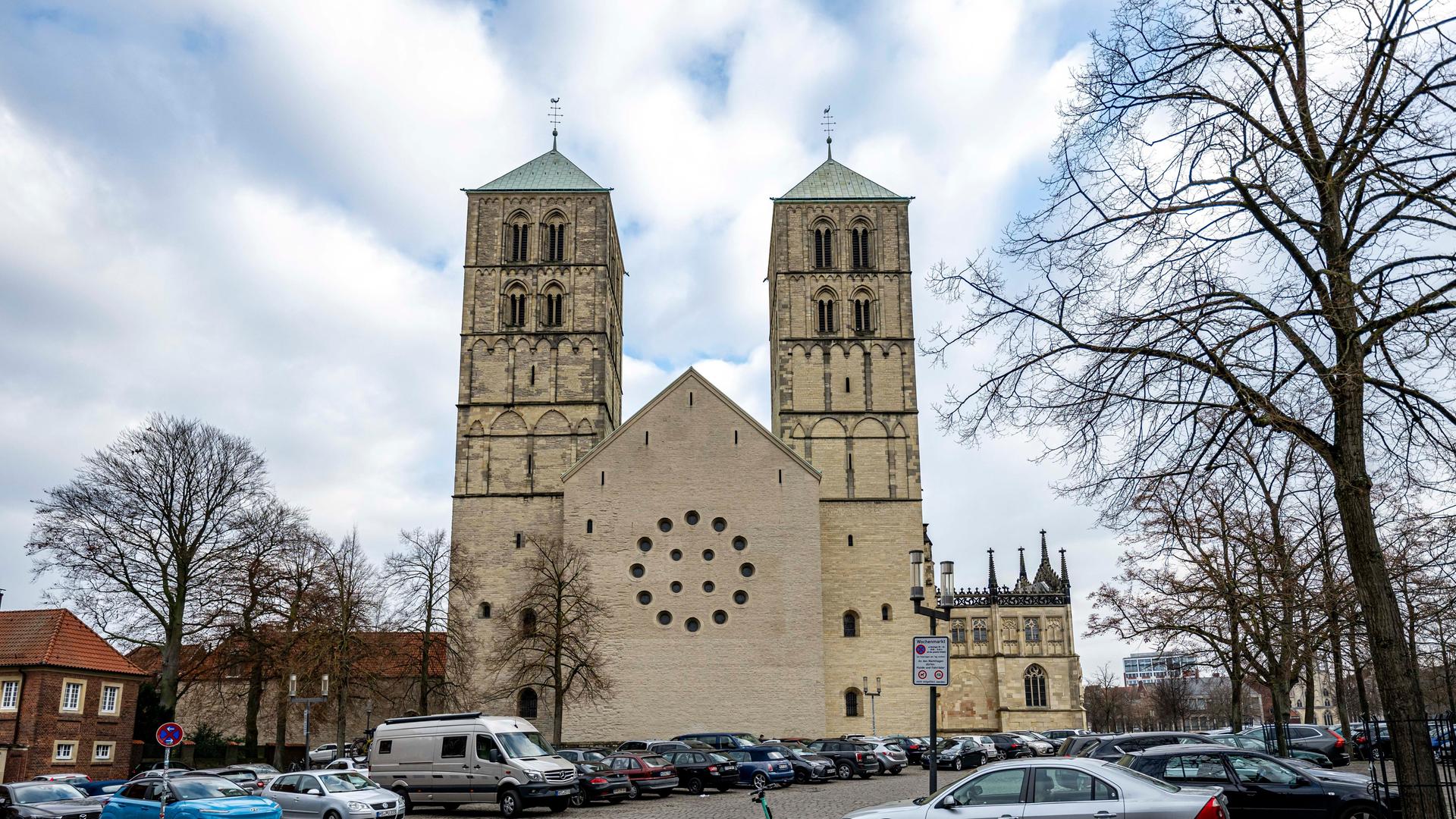 Blick auf das hellbraune Kirchengebäude vor einem großen Platz mit Pflastersteinen, auf dem viele Autos geparkt sind, und auf dem kahle Bäume stehen. Der Himmel darüber ist bewölkt. Blick auf das hellbraune Kirchengebäude vor einem großen Platz mit Pflastersteinen, auf dem viele Autos geparkt sind, und auf dem kahle Bäume stehen. Der Himmel darüber ist bewölkt.