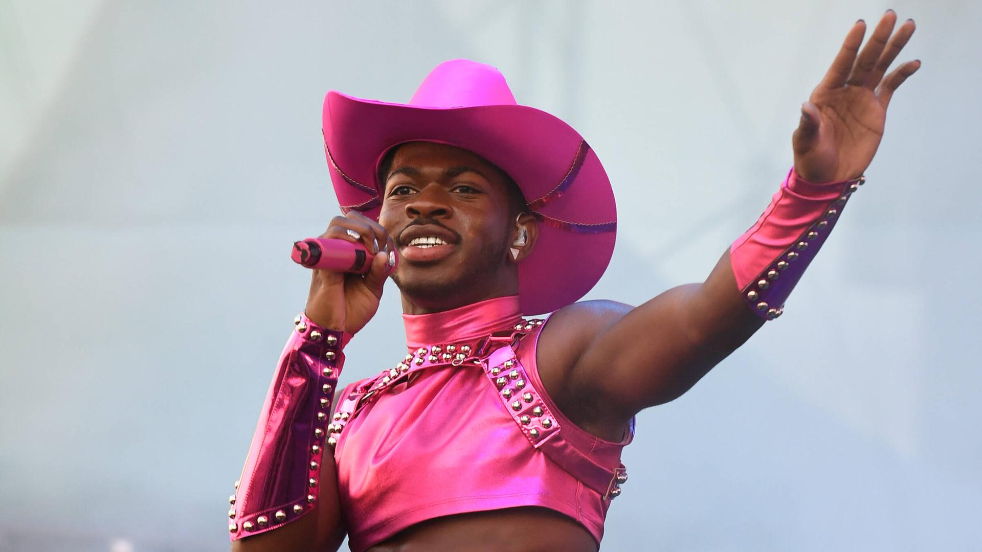 Lil Nas X steht auf einer Bühne. Er trägt einen pinken Cowboy-Hut, ein pinkes Top und ein pinkes Röckchen. In der Hand hält er ein pinkes Mikrofon. Lil Nas X steht auf einer Bühne. Er trägt einen pinken Cowboy-Hut, ein pinkes Top und ein pinkes Röckchen. In der Hand hält er ein pinkes Mikrofon.