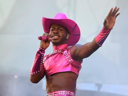 Lil Nas X steht auf einer Bühne. Er trägt einen pinken Cowboy-Hut, ein pinkes Top und ein pinkes Röckchen. In der Hand hält er ein pinkes Mikrofon.