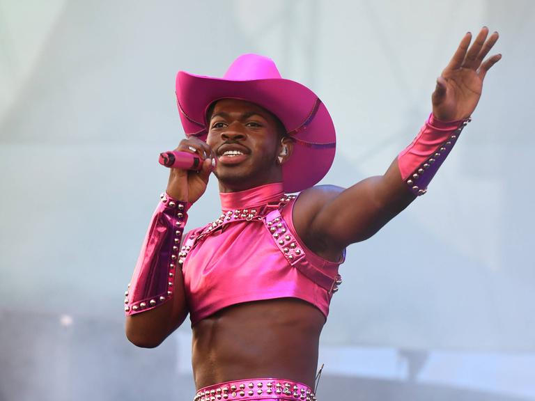 Lil Nas X steht auf einer Bühne. Er trägt einen pinken Cowboy-Hut, ein pinkes Top und ein pinkes Röckchen. In der Hand hält er ein pinkes Mikrofon.