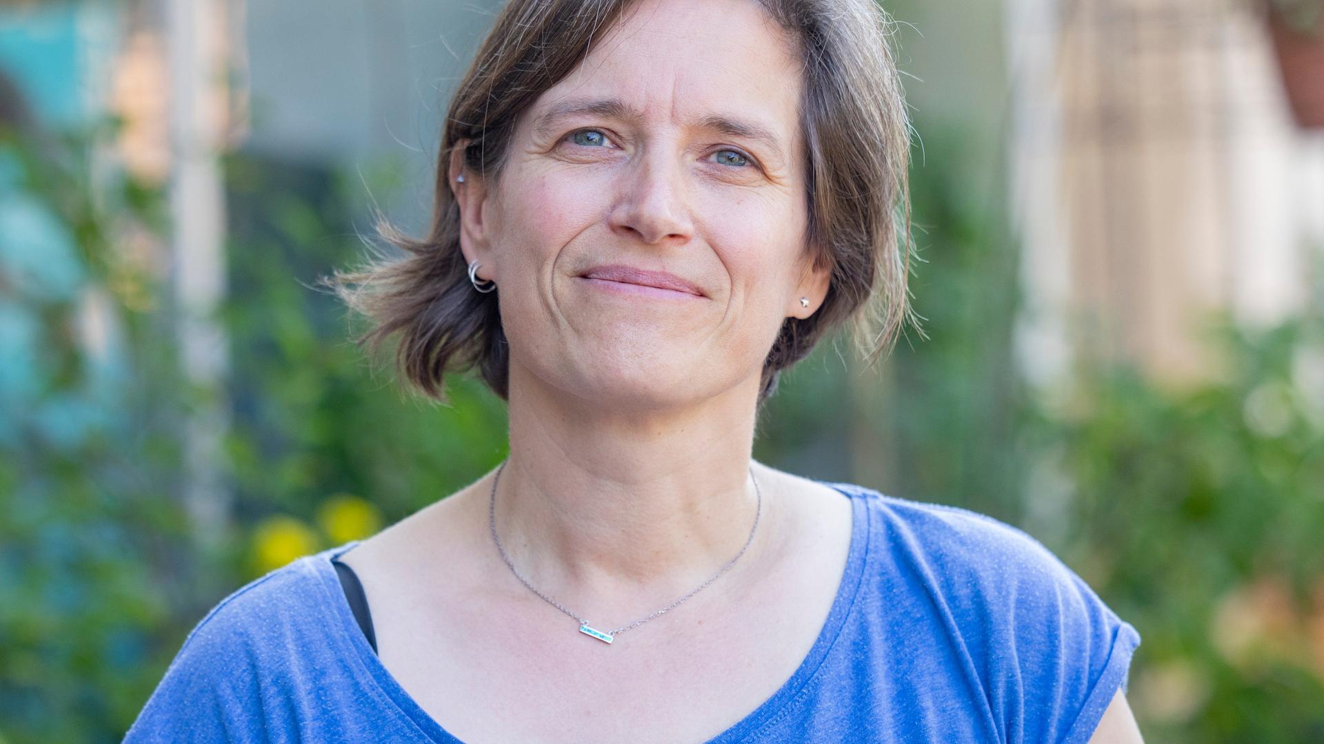 Die Traumatherapeutin Verena Fink steht vor einem verschwommenen grünen Hintergrund und blickt den Betrachter an. Sie trägt ein blaues T-Shirt und eine Halskette und hat halblange braune Haare. Die Traumatherapeutin Verena Fink steht vor einem verschwommenen grünen Hintergrund und blickt den Betrachter an. Sie trägt ein blaues T-Shirt und eine Halskette und hat halblange braune Haare.