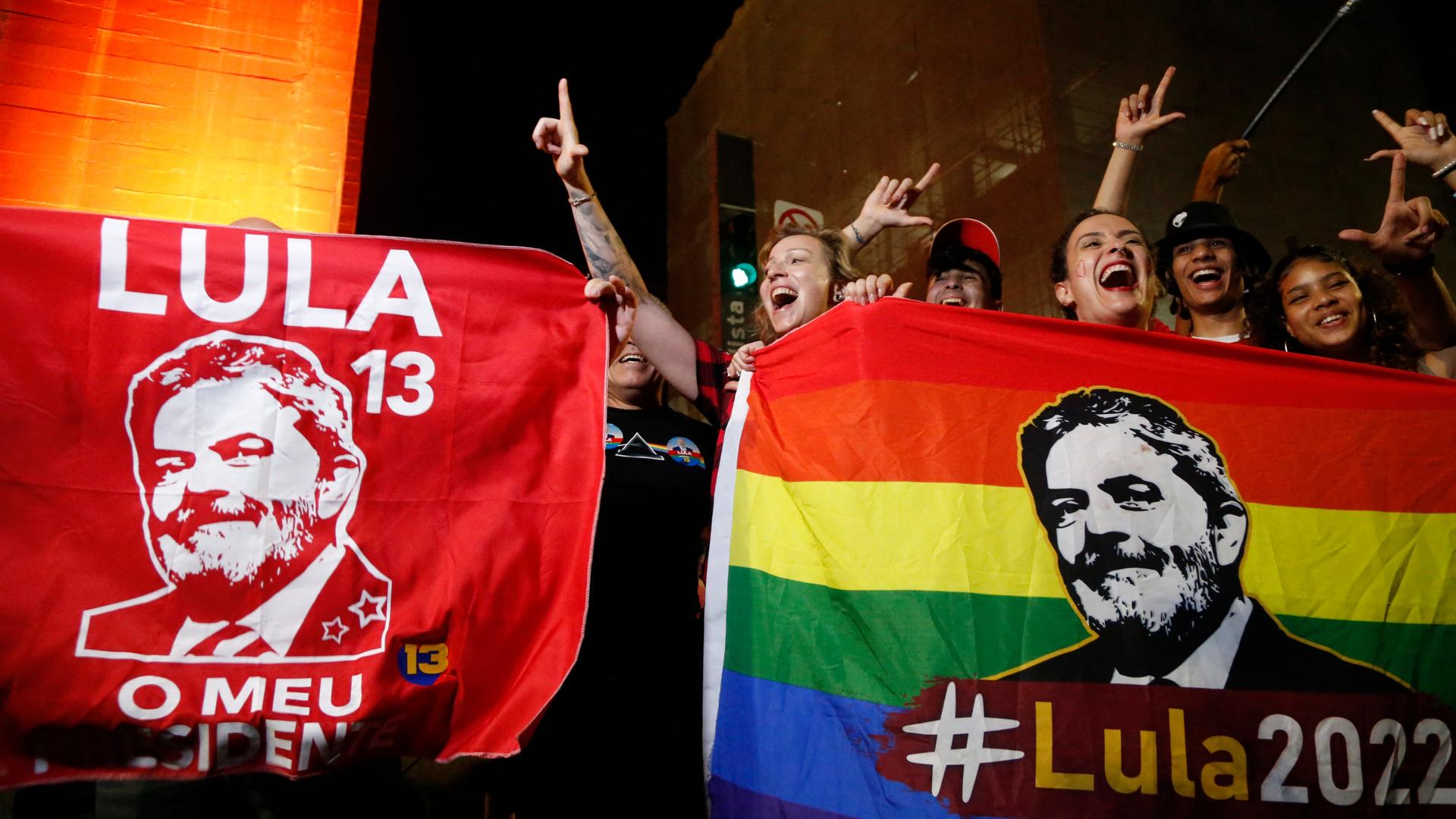 Zahlreiche Menschen feiern den Wahlsieg Lula da Silvas in Sao Paolo, Brasilien. Es ist dunkel, sie halten zwei bunte Flaggen mit dem Portrait Lulas hoch und sind fröhlich. Zahlreiche Menschen feiern den Wahlsieg Lula da Silvas in Sao Paolo, Brasilien. Es ist dunkel, sie halten zwei bunte Flaggen mit dem Portrait Lulas hoch und sind fröhlich.