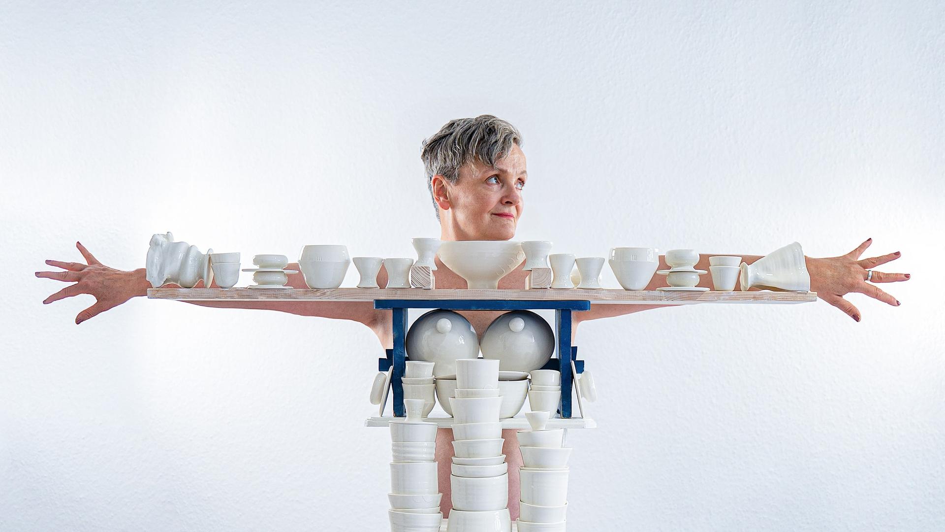 Die Künstlerin Uli Aigner steht hinter einem Gestell mit vielen weiße Porzellanobjekten aus ihrem Kunstprojekt "One Million", und breitet die Arme aus. Das Gestell mit den Porzellanteilen formt ihren Körper nach. Die Künstlerin Uli Aigner steht hinter einem Gestell mit vielen weiße Porzellanobjekten aus ihrem Kunstprojekt "One Million", und breitet die Arme aus. Das Gestell mit den Porzellanteilen formt ihren Körper nach.