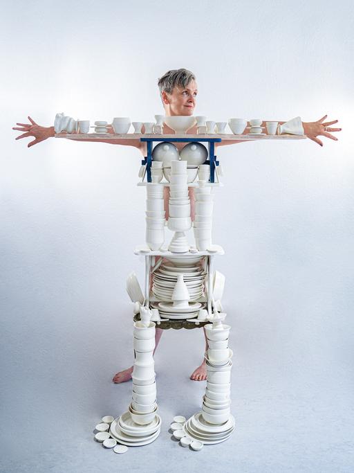 Die Künstlerin Uli Aigner steht hinter einem Gestell mit vielen weiße Porzellanobjekten aus ihrem Kunstprojekt "One Million", und breitet die Arme aus. Das Gestell mit den Porzellanteilen formt ihren Körper nach. 