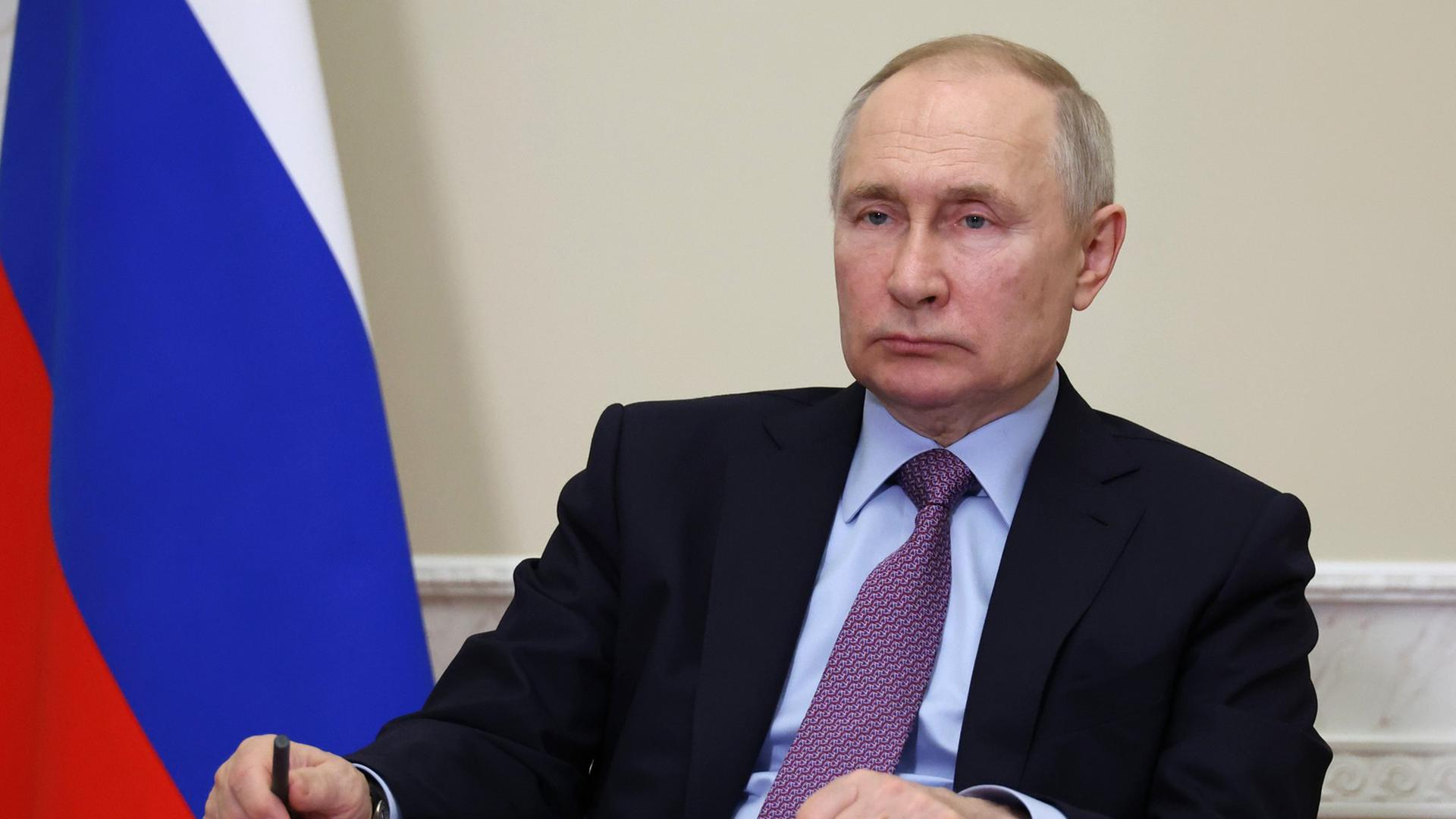 Wladimir Putin sitzt mit ernster Mine vor einer russischen Flagge und hat seine Hände auf einen Tisch gelegt.
