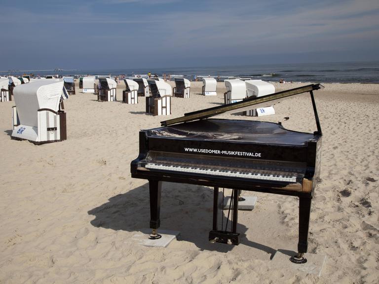 Wir sehen einen Klavier-Flügel am Strand, der vor vielen Strandkörben steht. Im Hintergrund das Meer. 