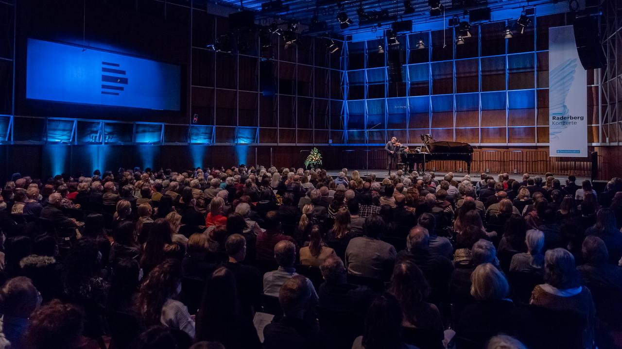 Ein Geiger und ein Pianist geben ein Konzert in einem voll besetzten, holzvertäfelten Saal, der blau beleuchtet ist. Ein Geiger und ein Pianist geben ein Konzert in einem voll besetzten, holzvertäfelten Saal, der blau beleuchtet ist.