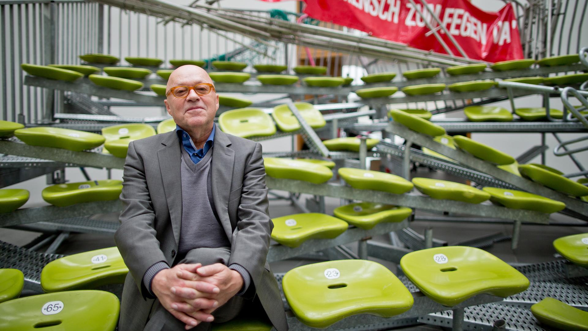 Der Künstler Olaf Metzel sitzt auf seinen kombinierten Installationen "Auf Wiedersehen" (1996/2015) und "In einer öffentlichen Halle ist nie ein Mensch zum Fegen da" (1996/2014) am 12.11.2015 im Neuen Museum in Nürnberg. Im Hintergrund ist ein Rotes Transparent mit einer weißen Aufschrift zu sehen sowie davor Sitzbankreihen mit grünfarbene Schalensitzen, die schief im Raum stehen und ineinander verwebt sind. Im Vordergrund sitzt Olaf Metzel, ein Mann mit Glatze. Er trägt einen dunkelgrauen Anzug über einem grauen Pullover und einem blauen Hemd.