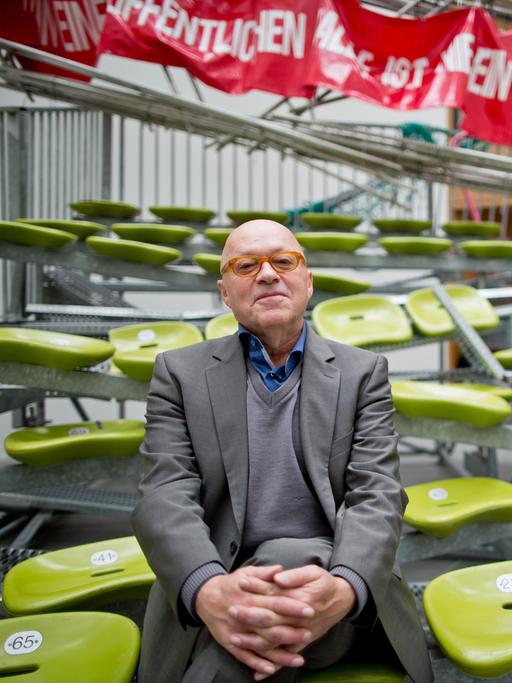 Der Künstler Olaf Metzel sitzt auf seinen kombinierten Installationen "Auf Wiedersehen" (1996/2015) und "In einer öffentlichen Halle ist nie ein Mensch zum Fegen da" (1996/2014) am 12.11.2015 im Neuen Museum in Nürnberg. Im Hintergrund ist ein Rotes Transparent mit einer weißen Aufschrift zu sehen sowie davor Sitzbankreihen mit grünfarbene Schalensitzen, die schief im Raum stehen und ineinander verwebt sind. Im Vordergrund sitzt Olaf Metzel, ein Mann mit Glatze. Er trägt einen dunkelgrauen Anzug über einem grauen Pullover und einem blauen Hemd.