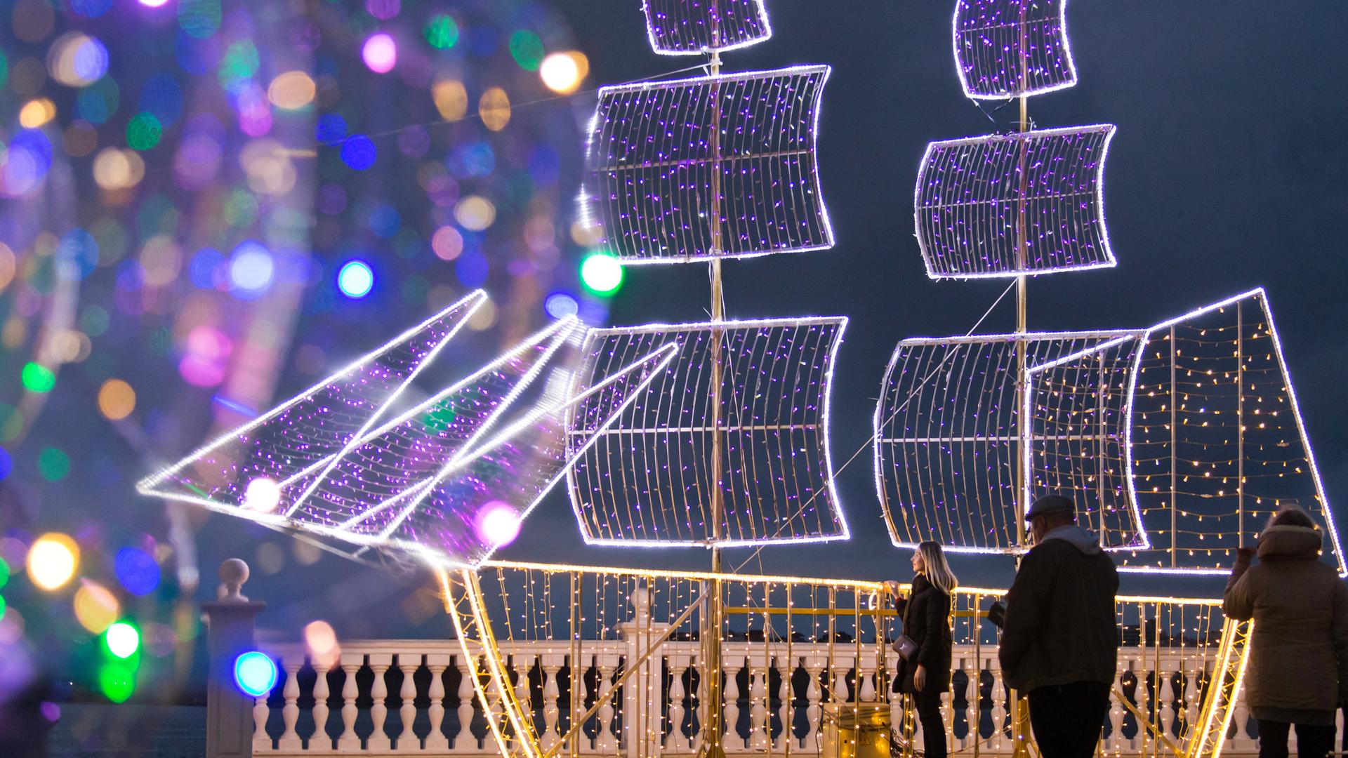 Bunte Lichter deuten am Meeresrand eine Schiffssilhouette an, die zu einer Weihnachtsbeleuchtung gehört.