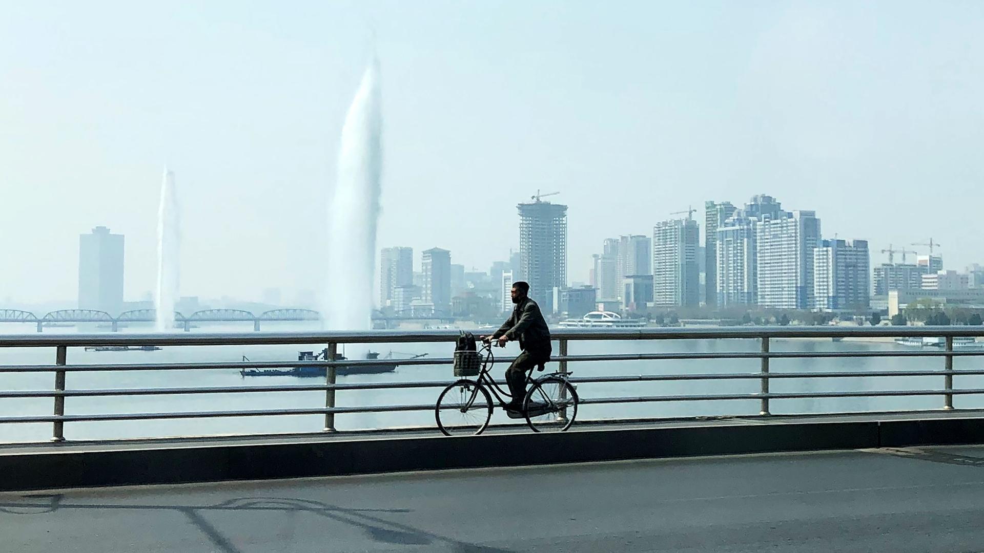 Ein Fahrradfahrer fährt über eine Brücke über den Taedong-Fluss in Pjöngjang. Im Hintergrund sind Hochhäuser zu sehen. Ein Fahrradfahrer fährt über eine Brücke über den Taedong-Fluss in Pjöngjang. Im Hintergrund sind Hochhäuser zu sehen.