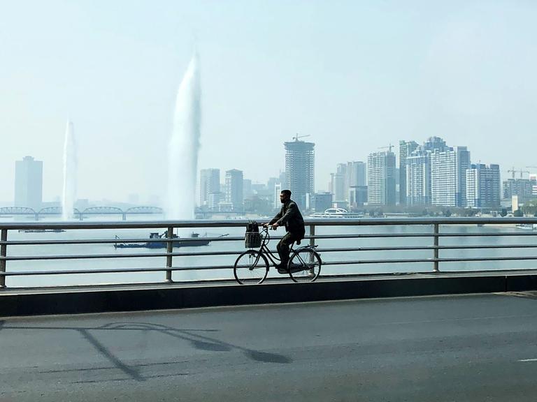 Ein Fahrradfahrer fährt über eine Brücke über den Taedong-Fluss in Pjöngjang. Im Hintergrund sind Hochhäuser zu sehen.